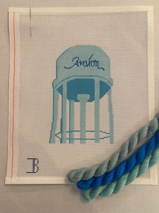A needlepoint canvas with a depiction of a water tower labeled 'Avalon' alongside two skeins of blue and green thread.