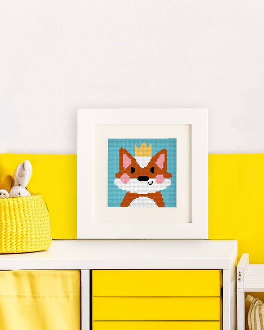 Framed illustration of a needlepoint fox wearing a crown on a yellow shelf with a white background
