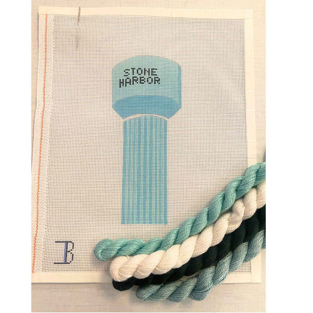 A needlepoint canvas with a depiction of a water tower labeled 'STONE HARBOR' in blue, surrounded by a frame, with additional colors of thread displayed in black and white.