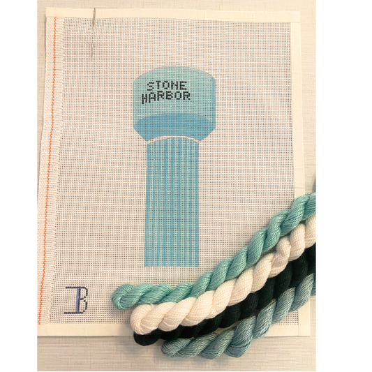 A needlepoint canvas with a depiction of a water tower labeled 'STONE HARBOR' in blue, surrounded by a frame, with additional colors of thread displayed in black and white.